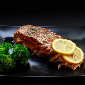 Grilled salmon on a black plate with two slices of lemon and broccoli.