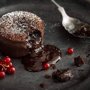 A single serving of warm chocolate lava cake sprinkled with powdered sugar with a bite taken out. The molten chocolate center spills out through the hole in the cakey wall. Red currant berries are the garnish for this indulgent dessert on a vintage metal plate.