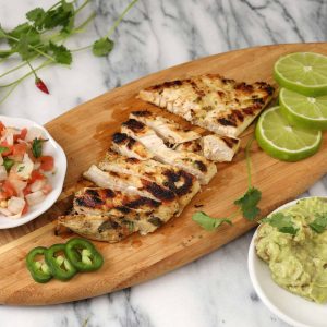 A high angle close up horizontal photograph of an oval wooden cutting board with sliced pieces of grilled chicken breast, limes, jalapeño slices and a bowl of pico de Gallo. A small white bowl of guacamole sits in the foreground.